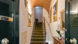 Una escalera conduce a una plataforma con techo de madera, una puerta negra con una placa y un mueble de madera con un jarrón de flores.