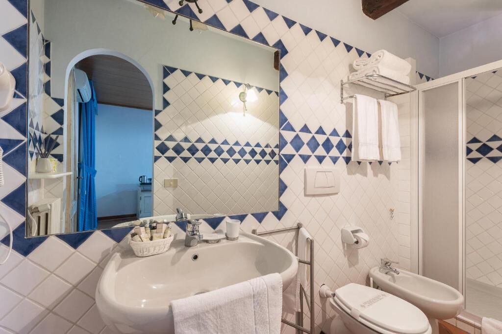 Une salle de bain avec un lavabo, des toilettes et une douche. Un miroir est placé au-dessus du lavabo et un porte-serviettes au-dessus des toilettes. Les murs sont recouverts de carreaux disposés selon un motif géométrique.