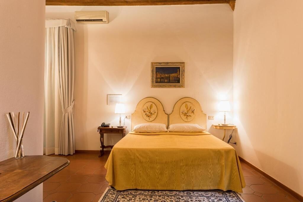 Un dormitorio con cama, dos mesitas de noche, lámpara, un cuadro en la pared y cortina.