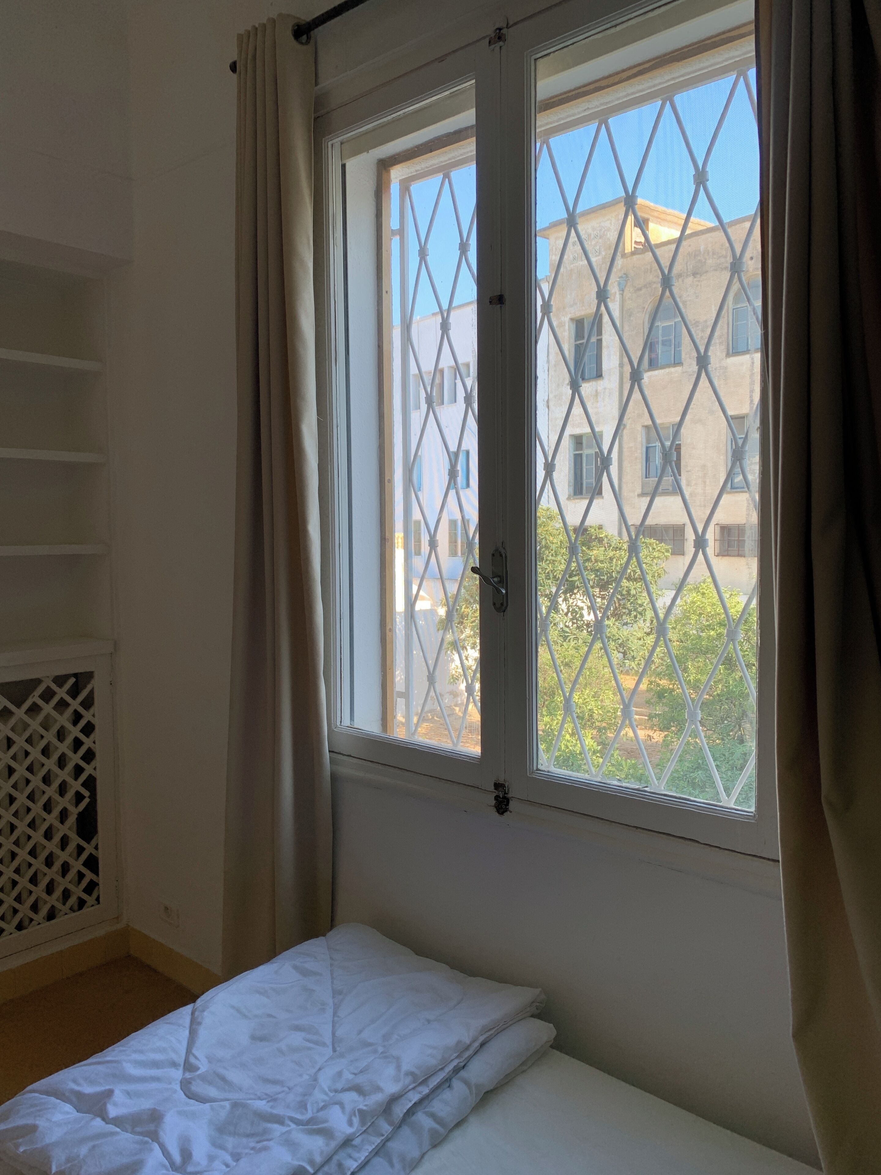 A room with a bed, a window with a metal grid, and a view of buildings outside.