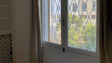 A room with a bed, a window with a metal grid, and a view of buildings outside.