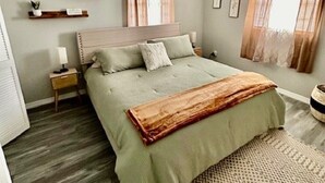 Un dormitorio con cama, mesitas de noche, un mueble auxiliar, una ventana con cortinas y una estantería con plantas.