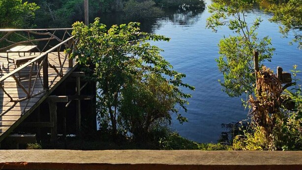 A wooden deck overlooking a calm river, surrounded by lush greenery and a thatched roof structure. - Pousada Cabana Nativa Paraíso (Careiro)