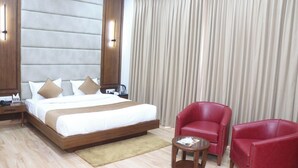 Quarto de hotel com uma cama grande, duas cadeiras vermelhas, uma mesa pequena e uma cortina.