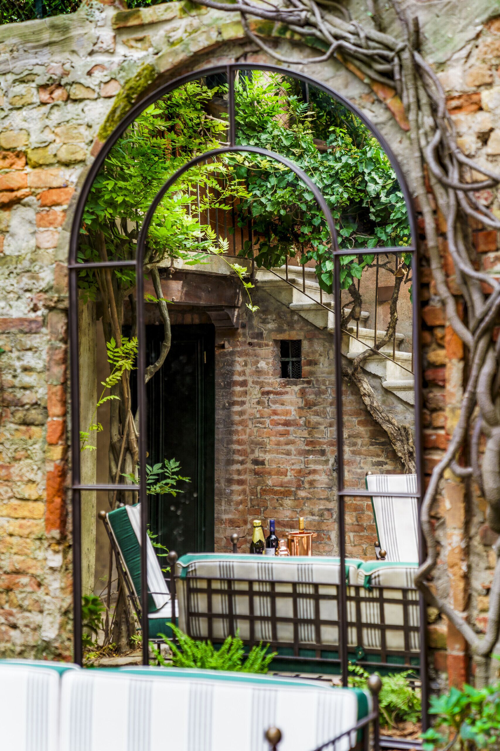 Un espace extérieur aménagé dans un style rustique, avec un mur en pierre, une arche en métal et un escalier menant à une petite pièce.