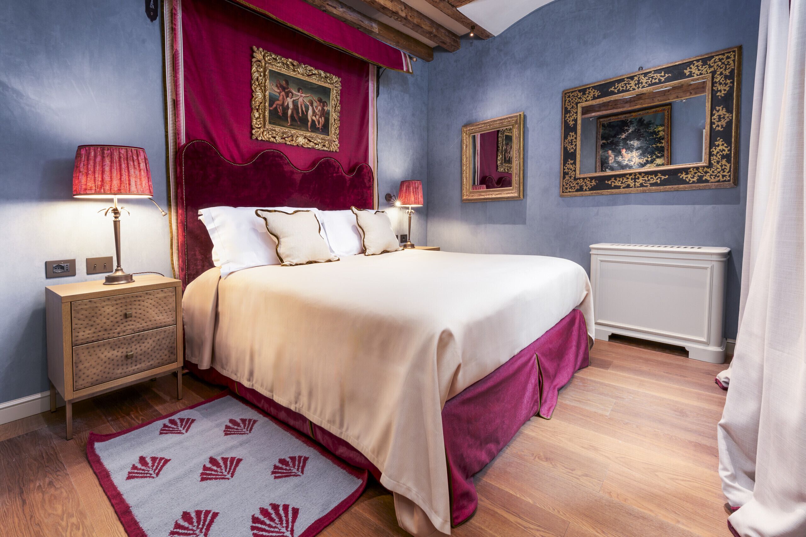 Une chambre à coucher avec un grand lit, deux tables de chevet, une commode, un tapis à motifs rouges et blancs, et un mur orné de deux miroirs encadrés et d’un tableau.