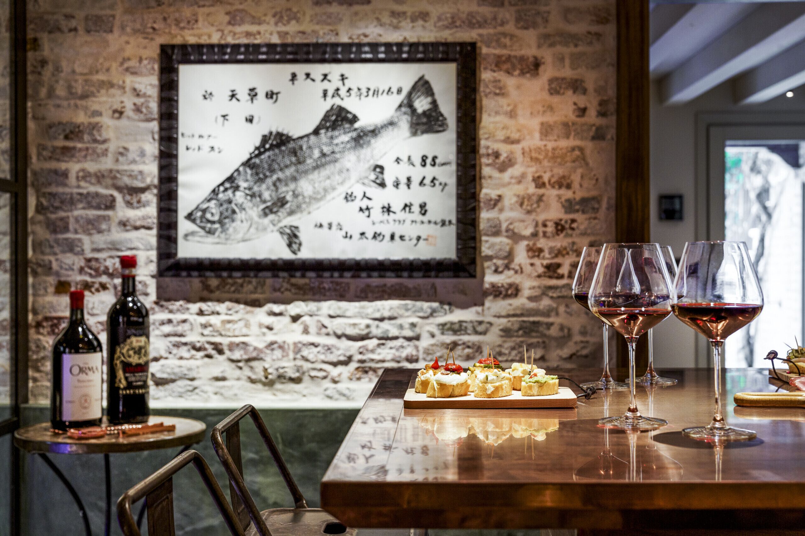 L’intérieur d’un restaurant avec un mur en briques, une illustration encadrée représentant un poisson, des bouteilles de vin, des verres et une petite table sur laquelle sont disposés des amuse-gueule.