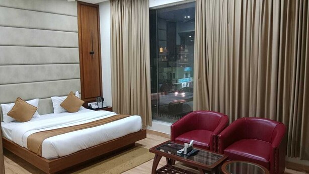 Une chambre d’hôtel avec un grand lit, deux fauteuils rouges, une table basse en verre et une vue sur la ville par une grande fenêtre.