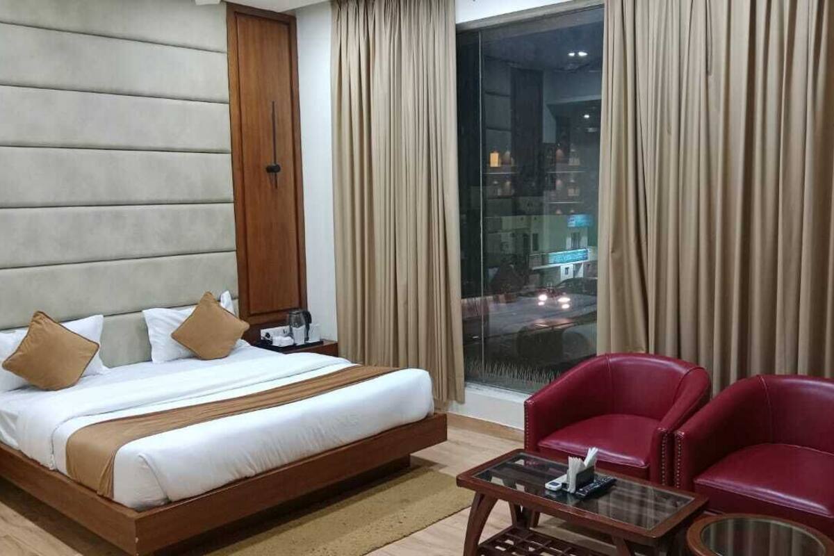 Une chambre d’hôtel avec un grand lit, deux fauteuils rouges, une table basse en verre et une vue sur la ville par une grande fenêtre.