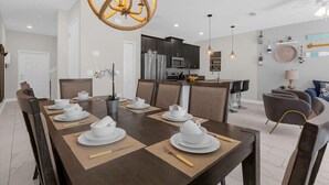 Un espace de salle à manger moderne, avec une table en bois foncé disposée pour six personnes, entourée de chaises. Un lustre original en forme de corde est suspendu au-dessus, et on aperçoit en arrière-plan une cuisine avec des meubles sombres et des appareils en acier inoxydable.