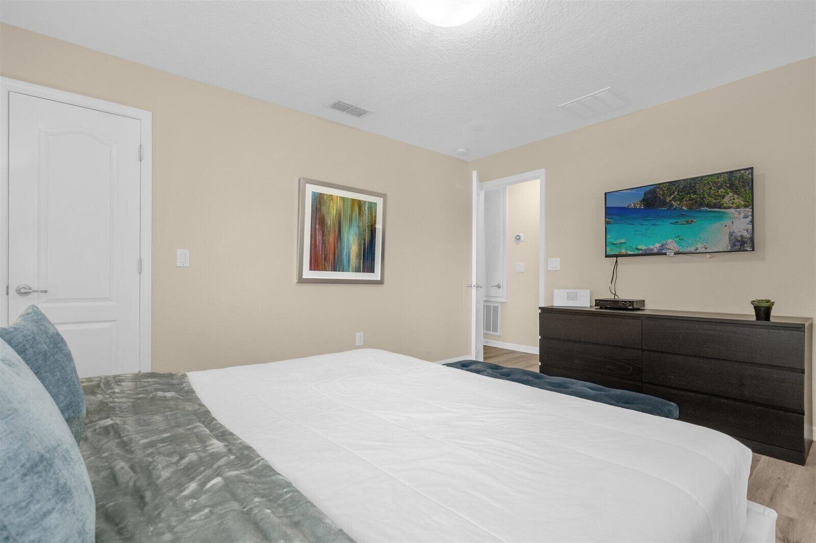 Une chambre à coucher avec un grand lit, une commode, un tableau représentant une plage et une porte.