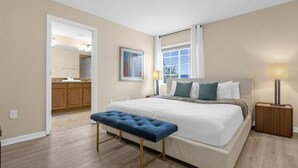 A bedroom with a large bed, a bench, a bedside table, a nightstand, a window with blinds, and a bathroom visible through an open door. - Cozy 5BD Home Paradise Palms Resort with Pool (Kissimmee)