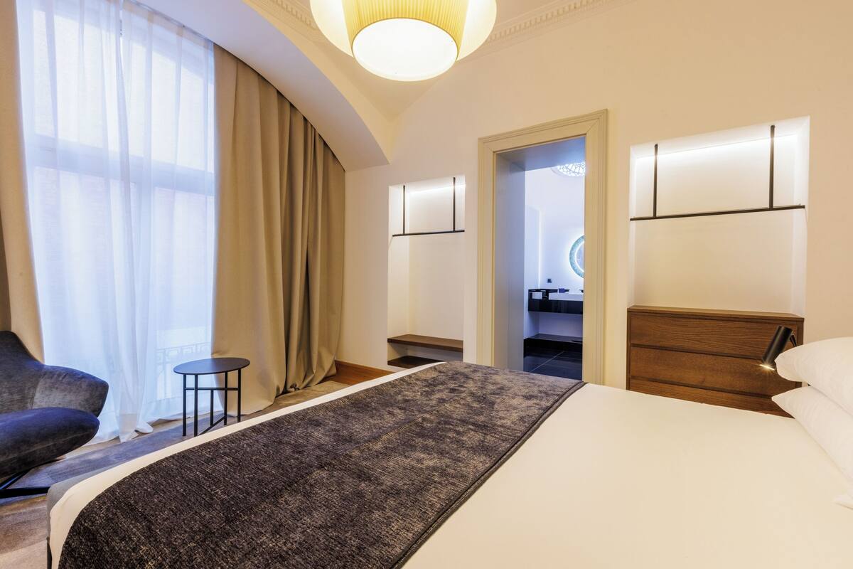 Habitación de hotel con una cama, una silla, una mesita, una mesita de noche y una puerta que da a un baño.