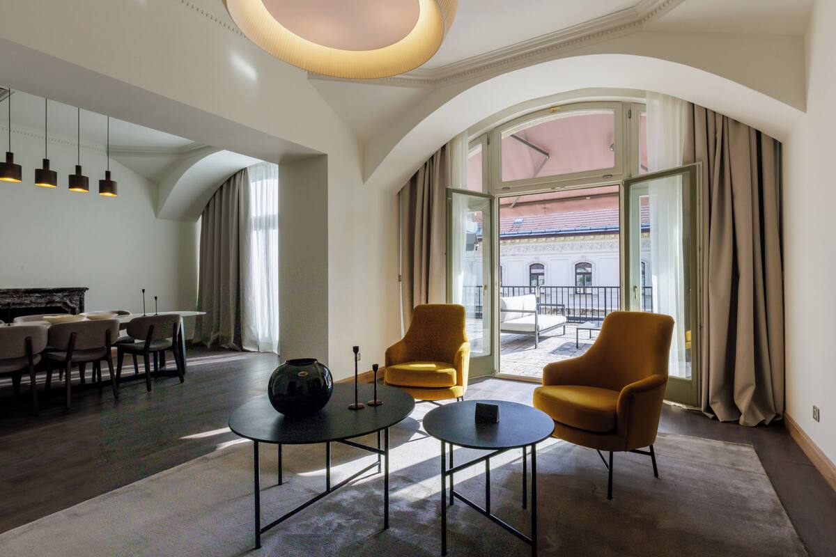 Una habitación de hotel moderna con zona de comedor, dos sillones, una mesita de centro y un ventanal con cortinas.