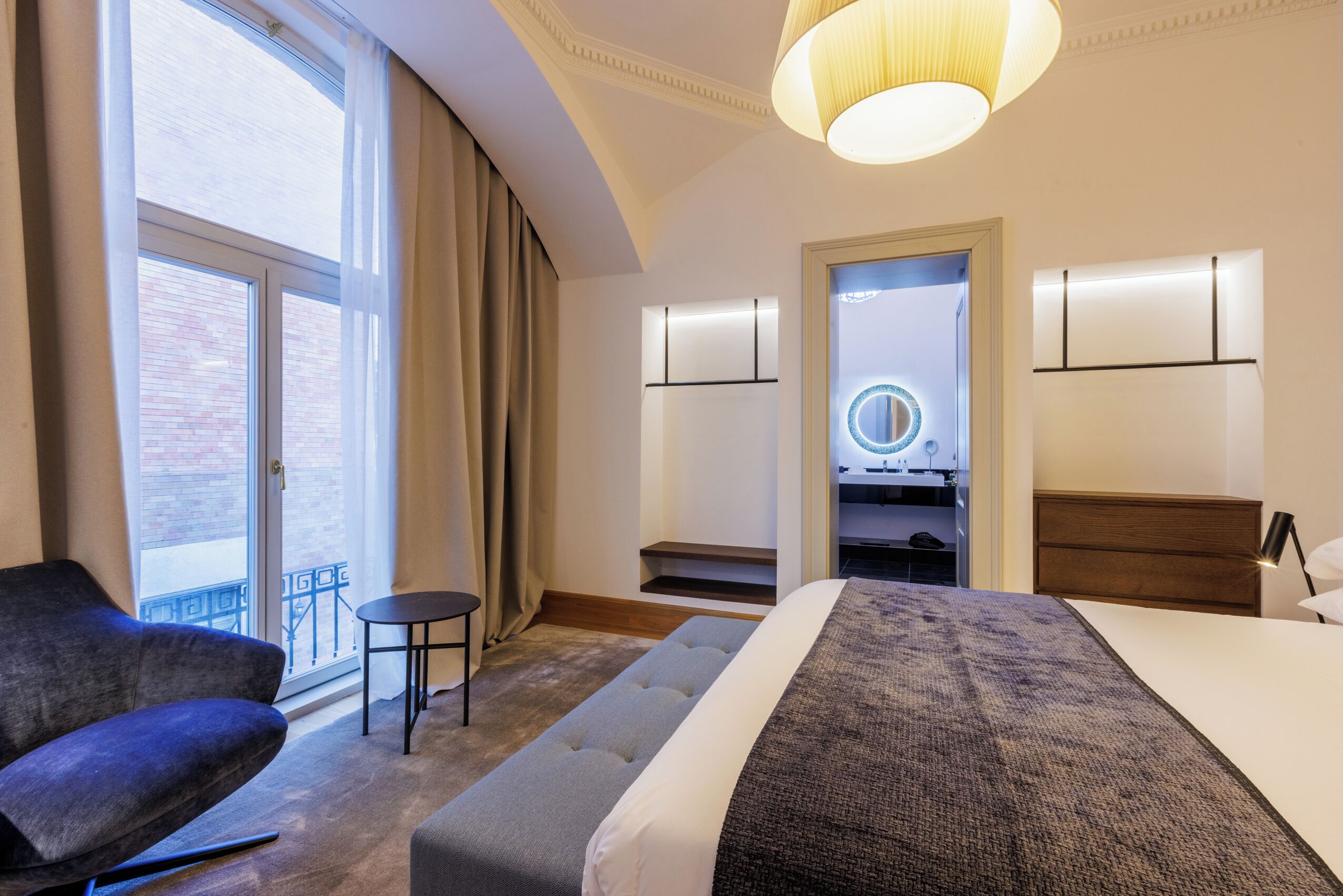 A hotel room with a bed, a chair, a small table, a mirror, and a view of the outside.