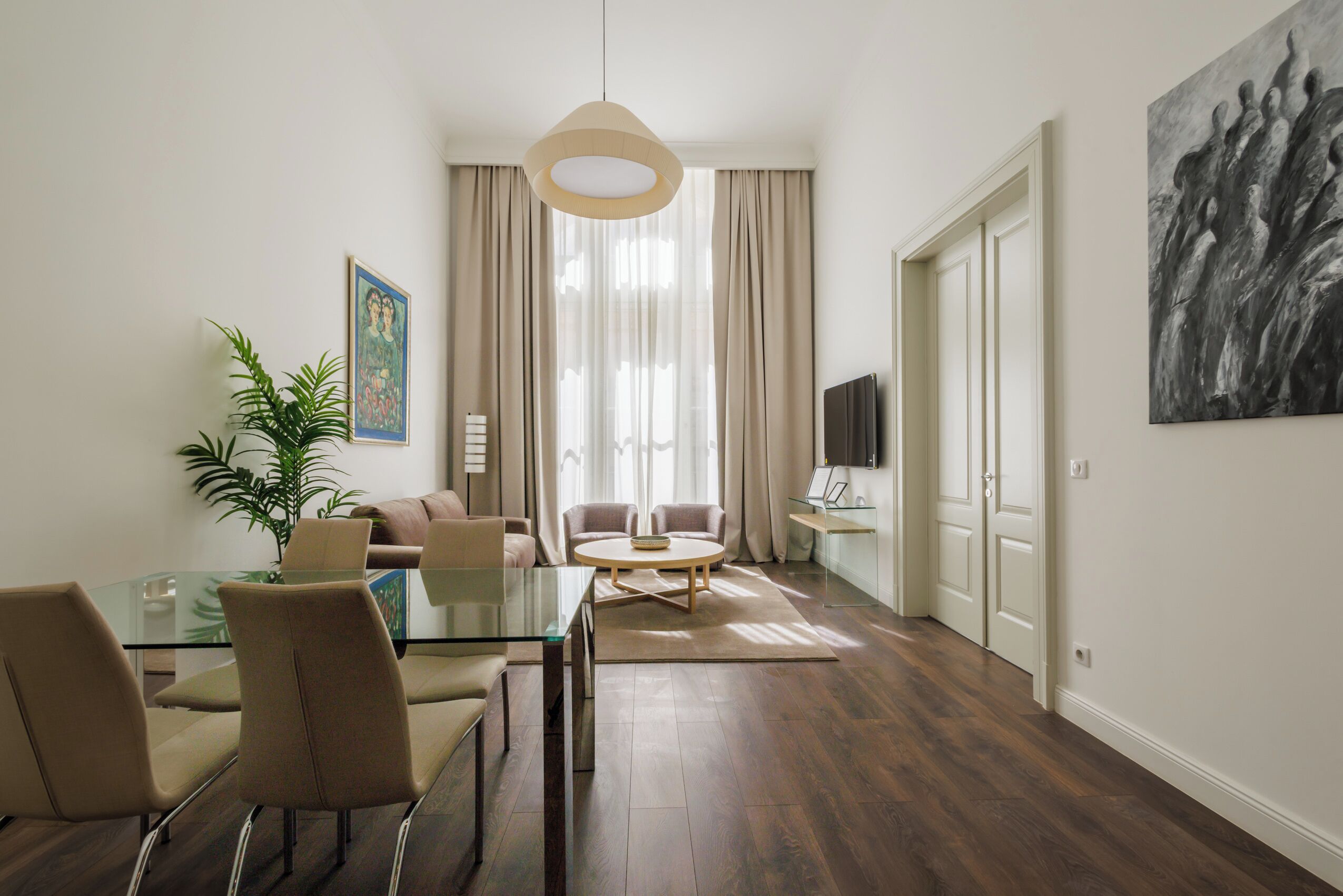 Een moderne woonkamer met een glazen eettafel, beige banken, een potplant en een groot abstract schilderij.