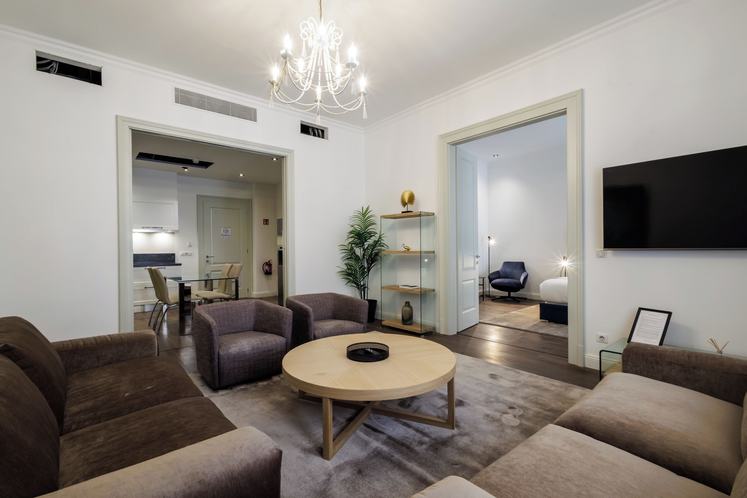 A modern living room with a round coffee table, two sofas, a chandelier, and a flat-screen TV mounted on the wall.