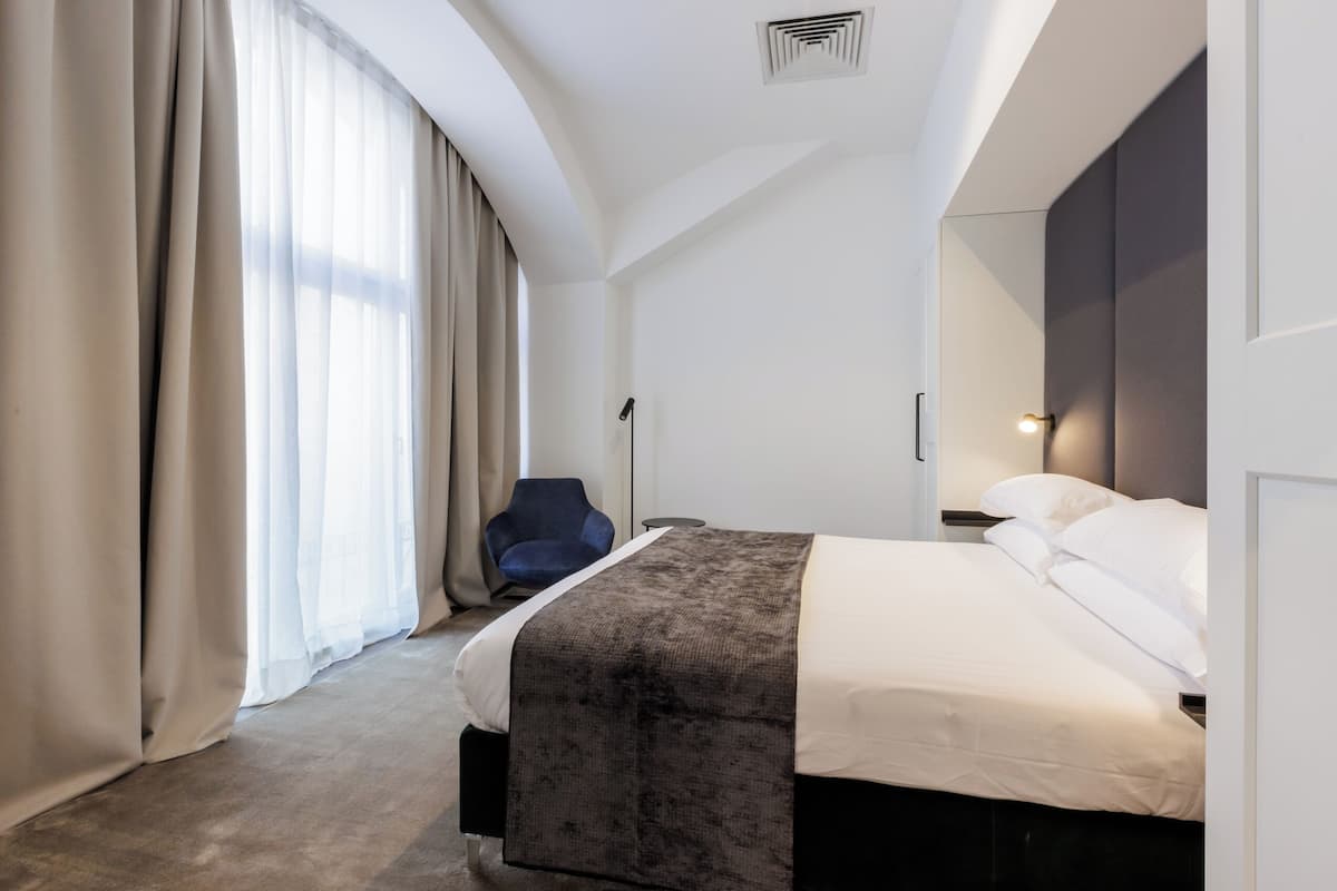 Una habitación de hotel moderna con una cama, una silla, un escritorio y un ventanal con cortinas.