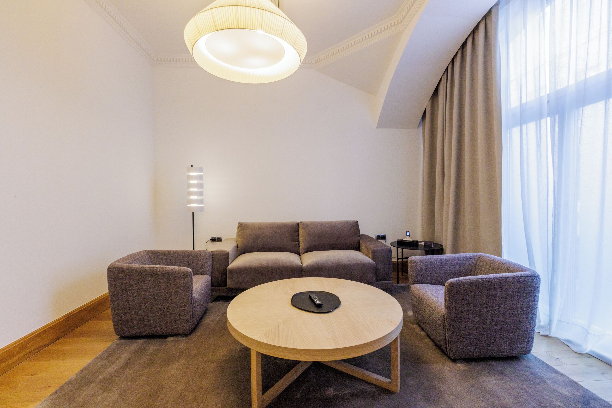 Una sala moderna con una mesa redonda, dos sillones, un sofá, una lámpara de pie y un ventanal con cortinas.