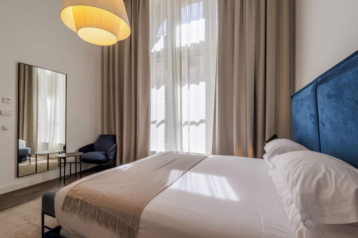Habitación de hotel con cama, una silla, una mesa, un espejo y ventana con cortinas.