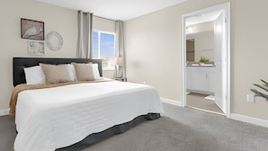 Ein Schlafzimmer mit Bett, Nachttisch, einem Fenster mit Vorhängen, einer Tür zu einem Badezimmer und Kunstwerken an der Wand.