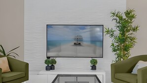 Moderni olohuone, jossa on seinälle asennettu litteänäyttöinen televisio, kaksi vihreää sohvaa koristetyynyineen, lasinen sohvapöytä ja ruukussa oleva kasvi.
