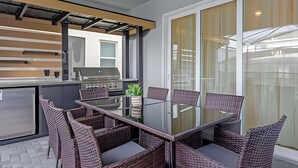 Ett modernt utomhusområde för måltider med ett glasbord och flätade stolar, en inbyggd grill och en skjutdörr i glas som leder ut till en balkong.