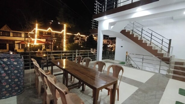 A dining area with a wooden table and chairs, a staircase, and a view of buildings in the background. - Hotel RISHI (Bhatwari)
