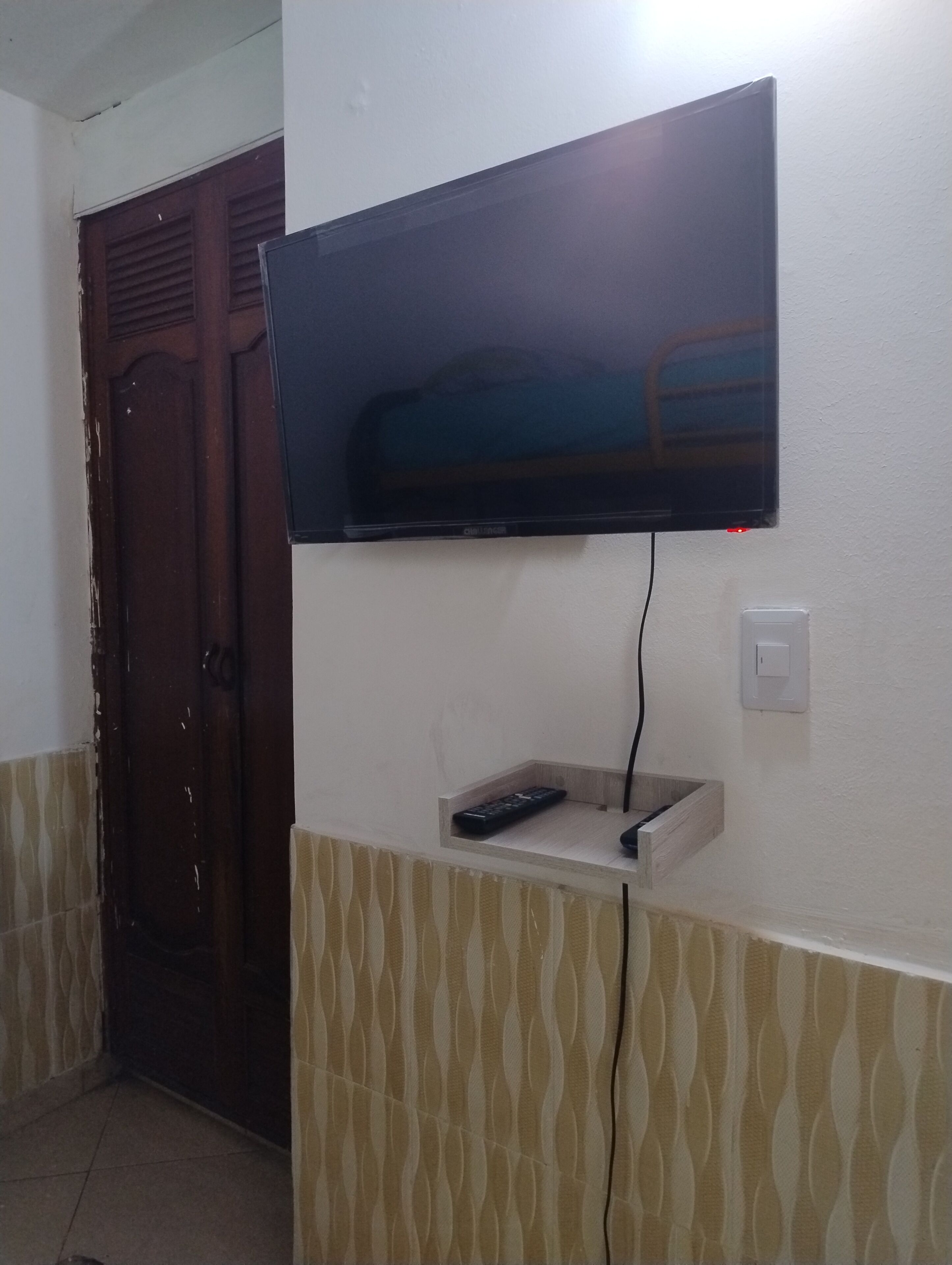 A television mounted on a wall with a remote control on a shelf below.