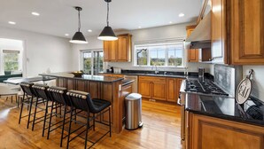 A modern kitchen with wooden cabinets, a central island with bar stools, a stainless steel trash can, and a gas stove. - Elegant Home w/7BRs, Game Room, Luxury Yard, Pool (Chester)
