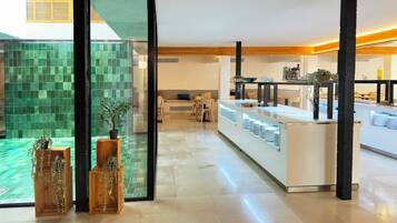 Una cocina moderna con una isla central, una zona de comedor con sofá y sillas, y vistas a un espacio exterior con azulejos verdes a través de un vidrio.