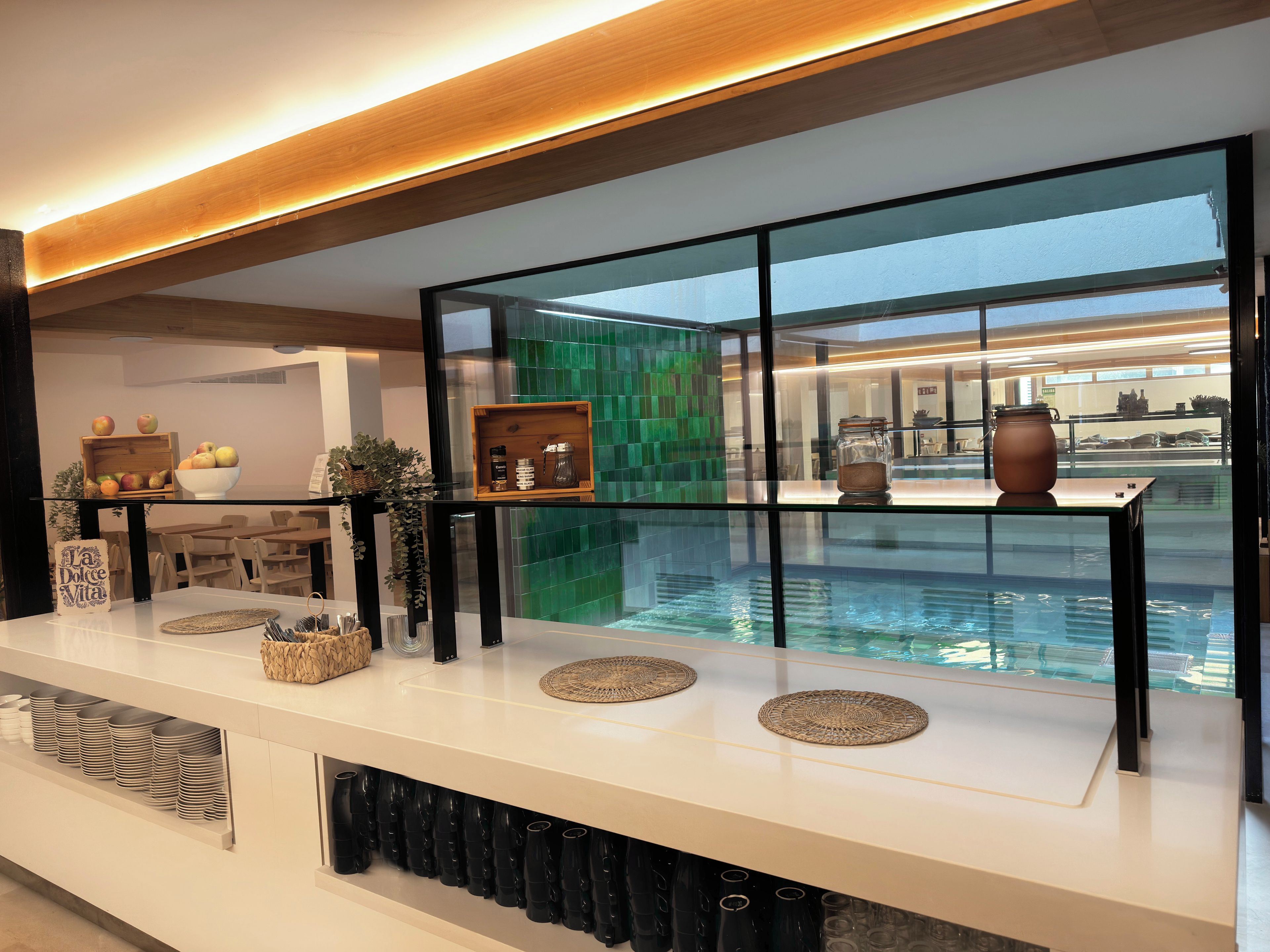 Vestíbulo moderno de hotel con separador de vidrio, vista a la piscina y área de recepción con mostrador y asientos de madera.