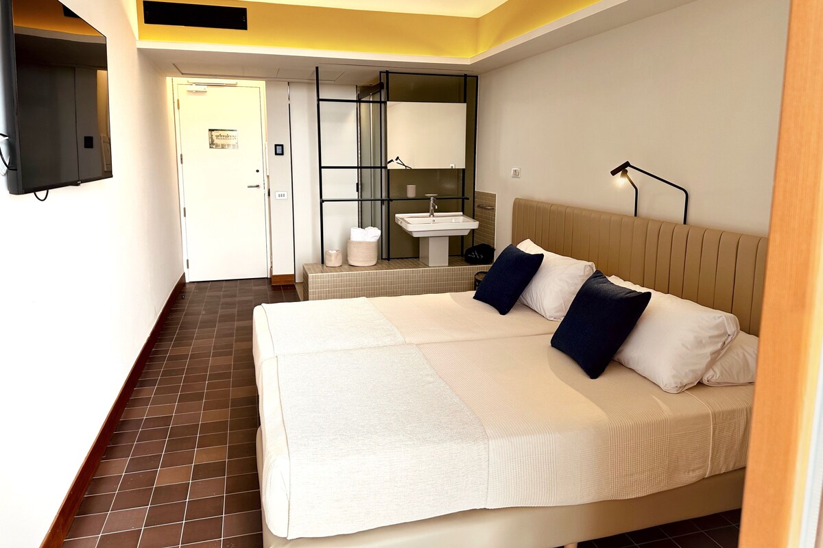 Habitación de hotel con una cama grande, una mesita de noche, un espejo, un televisor montado en la pared y un baño con lavamanos y espejo.