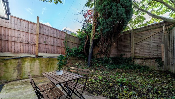 A wooden table and chairs set on a concrete patio with a wooden fence and greenery in the background. - The Cricklewood Apartment (London)