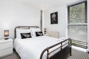 Ein modernes Schlafzimmer mit einem großen Bett, einem Nachttisch mit Lampe, einer Heizung und einem Fenster mit Jalousien.