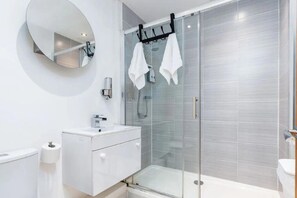 Ein modernes Badezimmer mit einer Duschkabine aus Glas, einem runden Spiegel und einem weißen Waschtisch mit Spüle und Handtuchhalter.