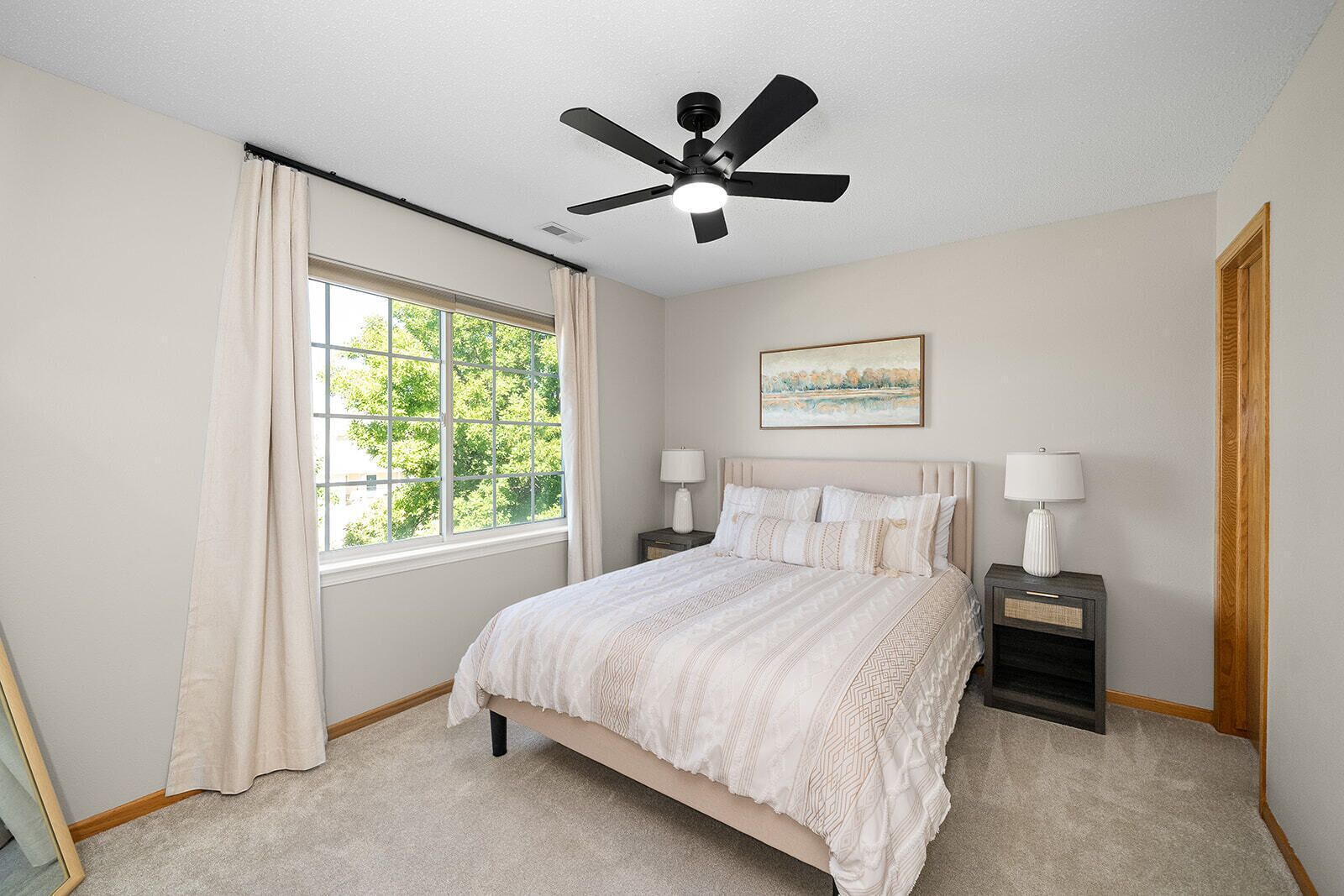 A bedroom with a bed, two bedside tables with lamps, a ceiling fan, a window with curtains, and a painting on the wall.