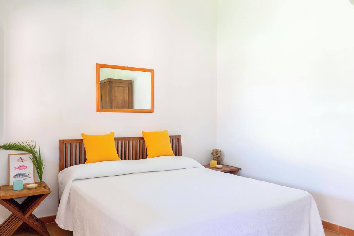 Un lit bien fait, avec une literie blanche et deux coussins jaune vif, une tête de lit en bois, une petite table de chevet avec une lampe et, au mur, un tableau représentant des poissons.
