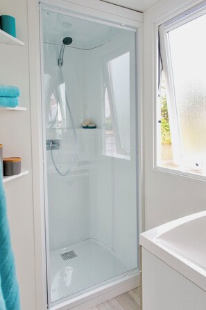 Un espace douche clos par des parois de verre, doté d’un pommeau de douche à main, d’une baignoire blanche et d’une fenêtre offrant une vue sur l’extérieur.