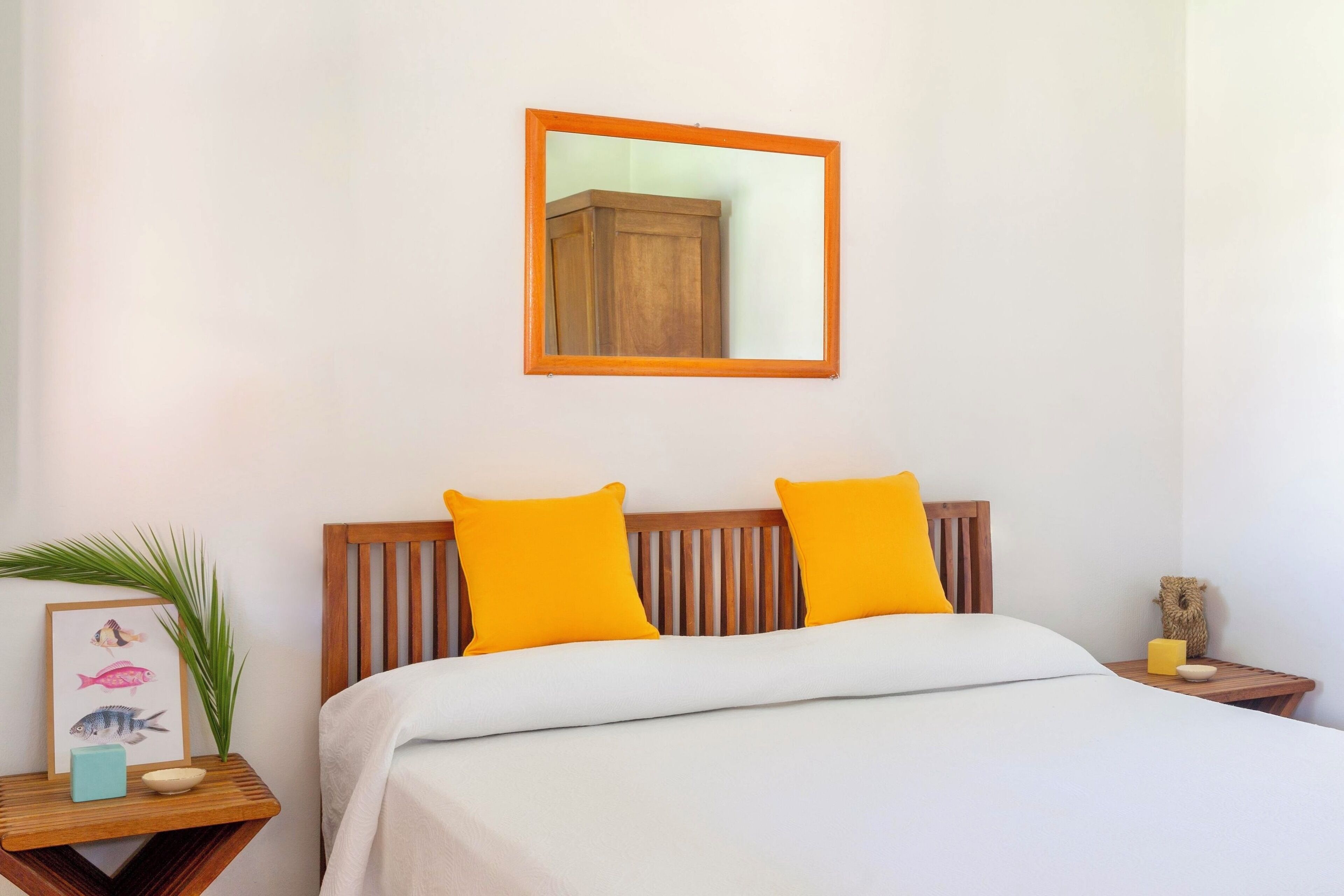 A bedroom with a wooden headboard, two bright yellow pillows, a white bedspread, a framed picture of fish, a small plant, and a nightstand with a candle and a small bowl.