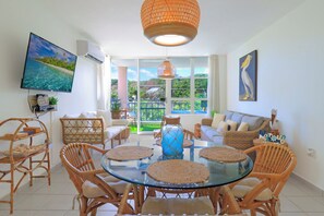 A bright living room with a glass dining table surrounded by wicker chairs, a large TV on the wall, and a balcony with a view of greenery. - The Escape House - condo in Ceiba (Ceiba)