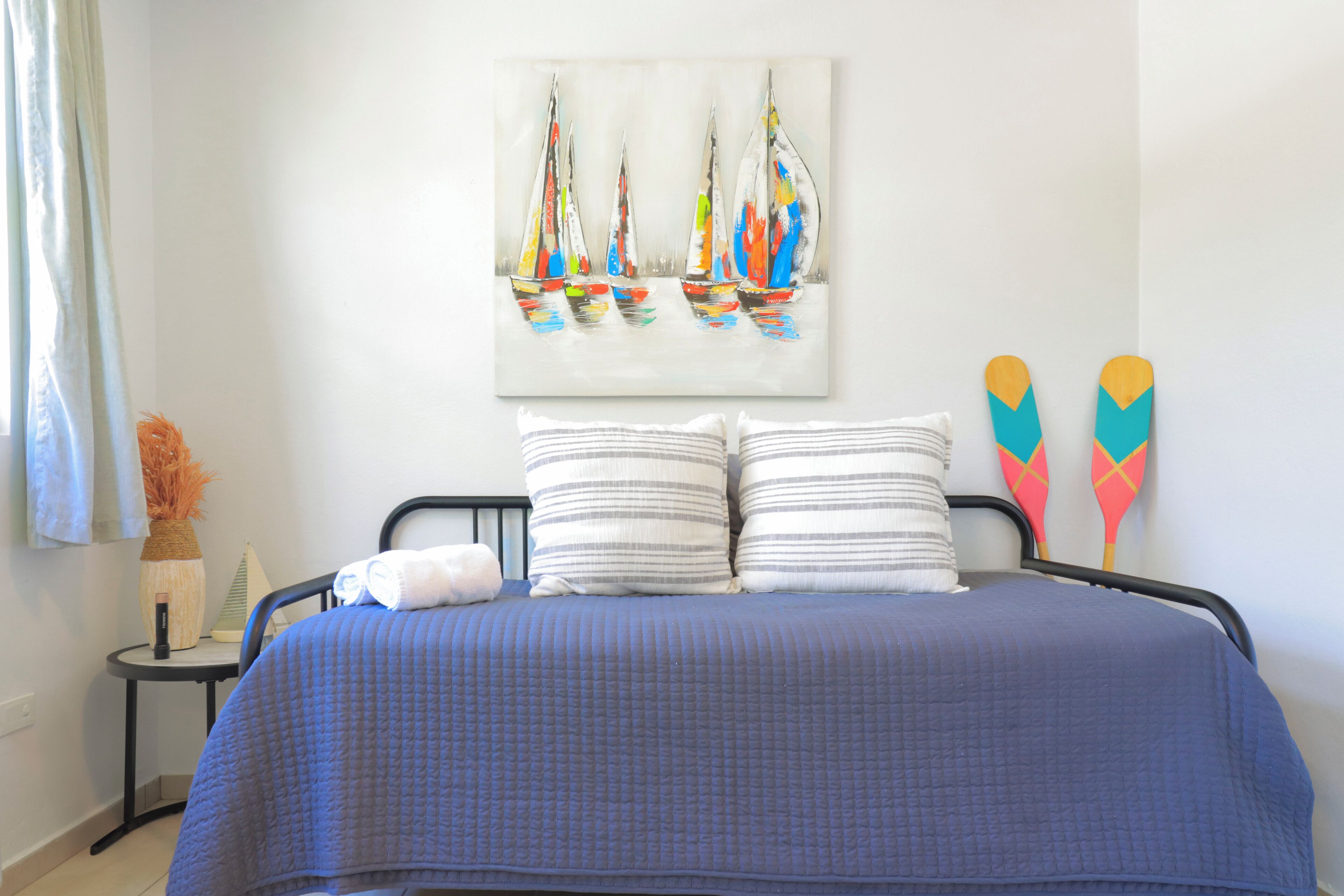 Un dormitorio con cama, almohadas, un cuadro de veleros, una mesita con un florero y una ventana con cortinas.