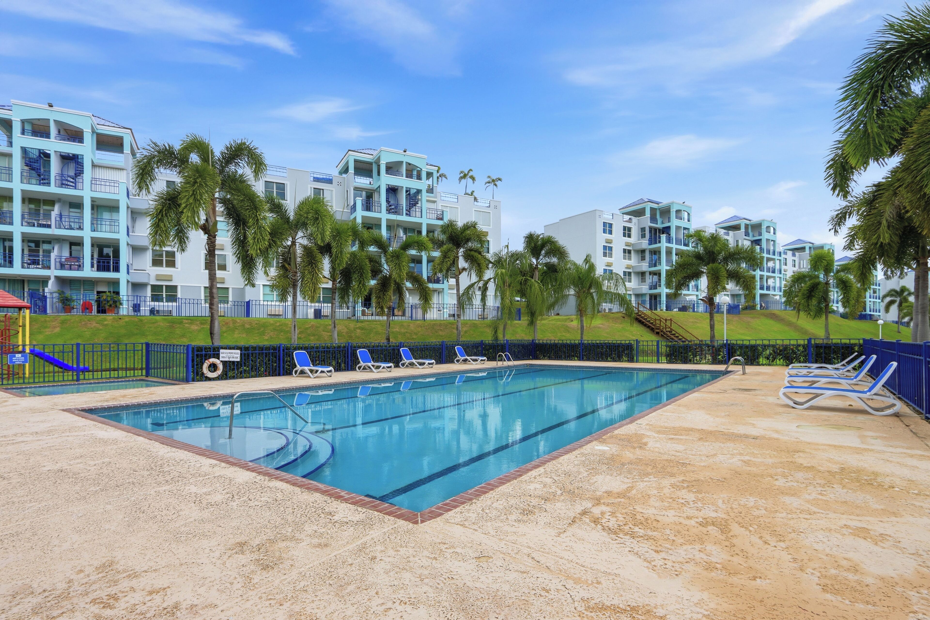 A swimming pool with lounge chairs, surrounded by palm trees and a building with multiple balconies.