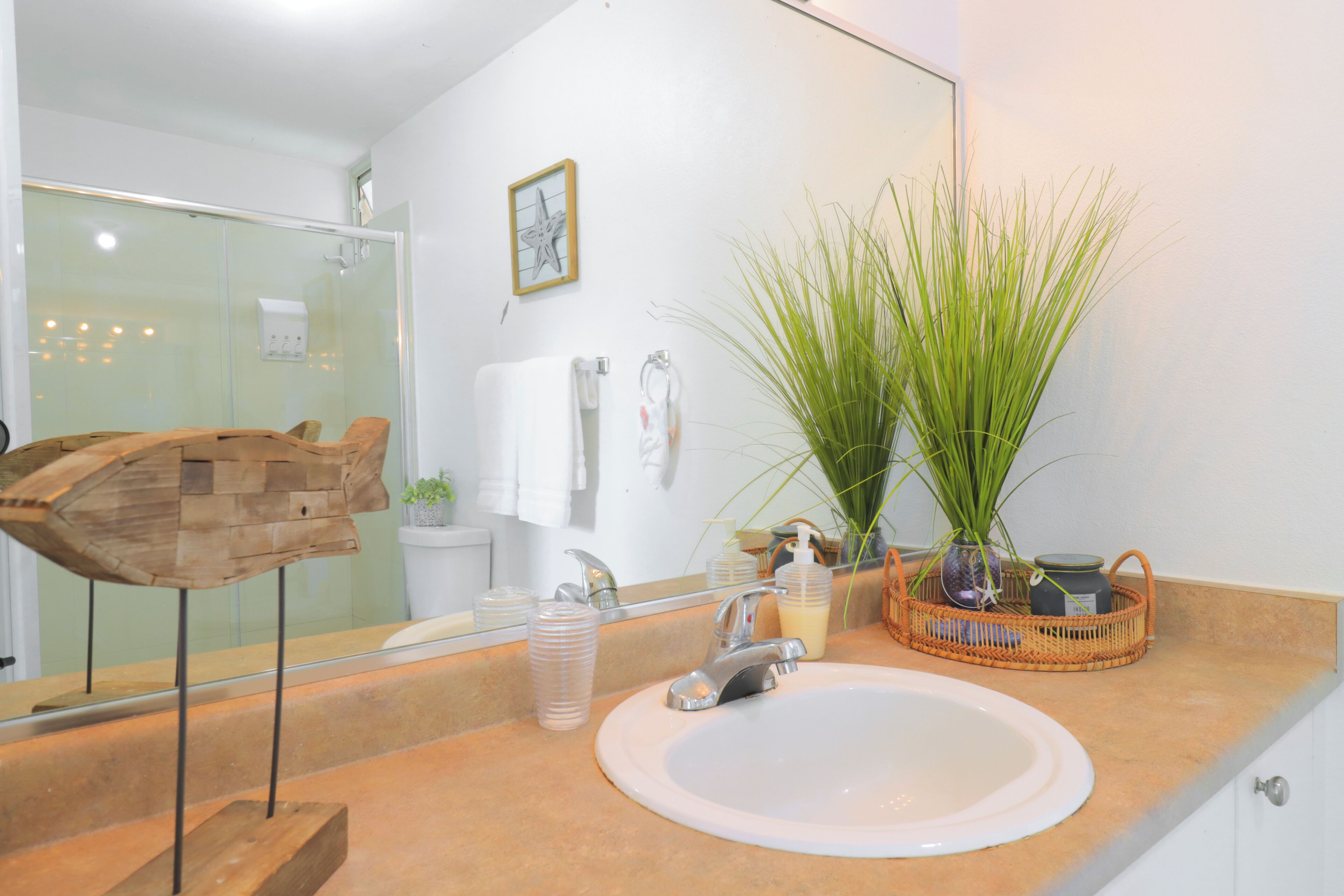 Un baño con lavabo blanco, espejo, escultura de madera, planta en maceta y zona de ducha.