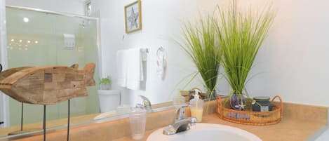 Un baño con lavabo blanco, espejo, escultura de madera, planta en maceta y zona de ducha.