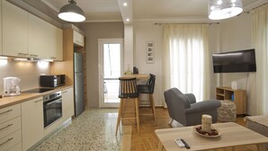 A modern kitchen with a built-in oven, microwave, and refrigerator. There is a dining area with a table and chairs, and a living room with a sofa and a TV. - Fabi's Place Luxurious 2BR, Fully Renovated Apt (Kipseli)