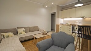 A modern living room with a sofa, a coffee table, and a grey armchair. A kitchen area is visible in the background with cabinets and a dining table. - Fabi's Place Luxurious 2BR, Fully Renovated Apt (Kipseli)