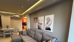 A modern living room with a grey sectional sofa, a dining table with chairs, and a kitchen area in the background. - SeaNfinity Spotless, 1BR Apt with Beach View (Nea Palatia)