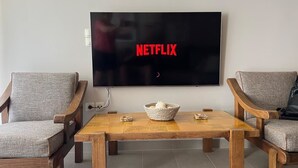 A living room with a large flat-screen TV mounted on the wall displaying the Netflix logo. There is a wooden coffee table with a woven basket in the center, flanked by two upholstered armchairs. - SeaNfinity Spotless, 1BR Apt with Beach View (Nea Palatia)