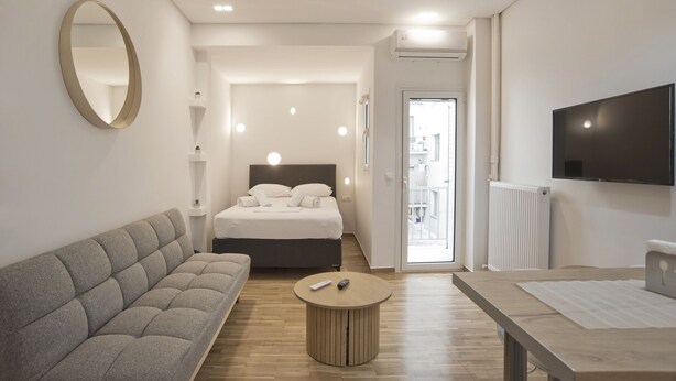 Una habitación de hotel moderna con un sofá, una mesa de centro redonda, una cama con cabecero, un televisor montado en la pared y un balcón visible a través de la puerta.